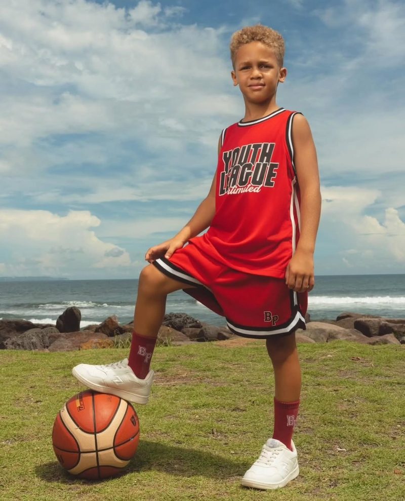 Keanu Tetteh in a red basketball kit with his foot on a ball by the sea